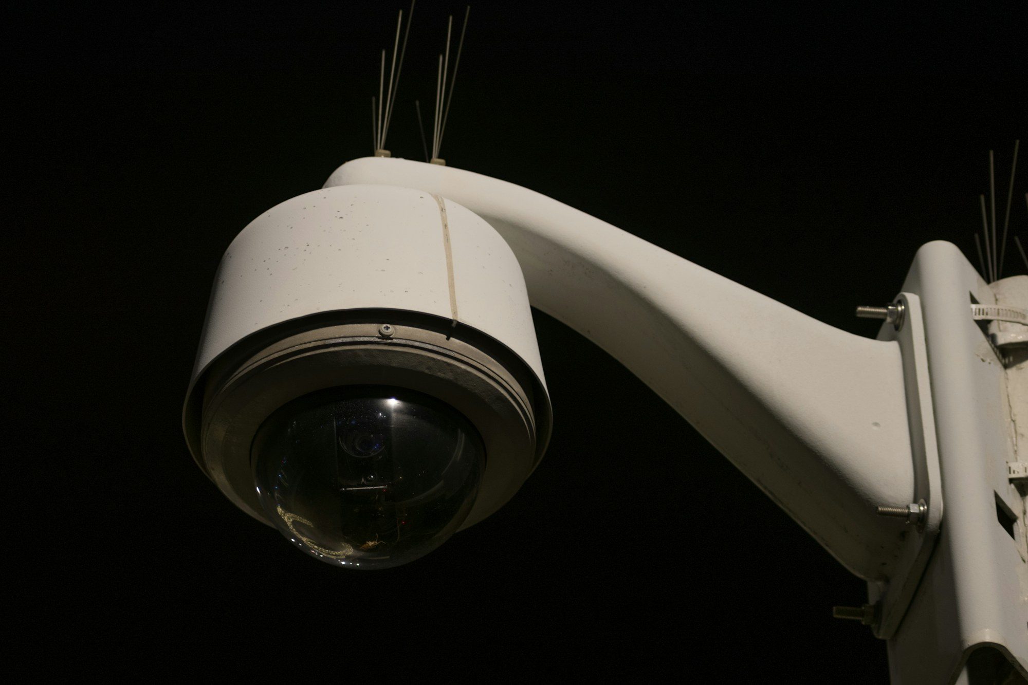 A pan-tilt-zoom dome camera on a wall arm, lit against a night sky